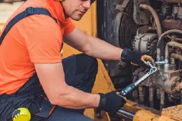 Mechanik w pomarańczowej koszulce naprawia układ hydrauliczny maszyny za pomocą klucza dynamometrycznego.